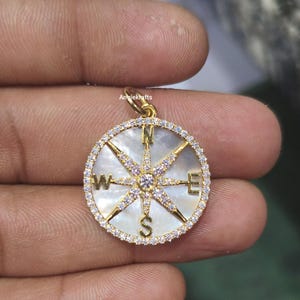 May include: Gold compass pendant with a white mother of pearl face and clear crystal accents. The compass points are marked with the letters N, S, E, and W.