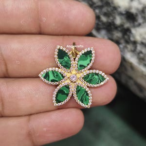 Puede incluir: Un colgante de flor de oro con piedra de malaquita verde y acentos de circonita cúbica transparente. El colgante está en una mano.