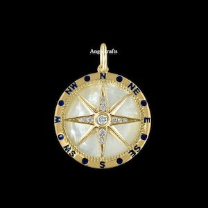 Puede incluir: Colgante de brújula de oro con una cara de nácar blanco y detalles en esmalte azul. La brújula presenta un diseño de estrella con diamantes transparentes.