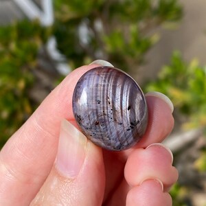 May include: A polished, oval-shaped gemstone with a mix of purple, gray, and brown hues. The stone has a glossy finish and displays a striped pattern. Small dark spots are visible on the surface. The gemstone is held in a hand.