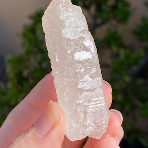 May include: A clear, raw quartz crystal with a rough, textured surface. The crystal is held in a hand, showcasing its natural, unpolished form. The quartz has a translucent appearance, reflecting light and revealing internal structures.