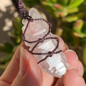May include: A clear, faceted quartz crystal point pendant wrapped in dark brown cord. The crystal features a natural, irregular shape with a translucent appearance and subtle iridescent highlights. A knotted cord loop extends from the top.
