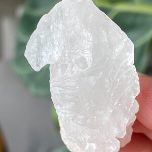 May include: A clear, raw quartz crystal with a rough, textured surface. The crystal is held in a hand, with a blurred green background. The quartz has a natural, irregular shape and is translucent.