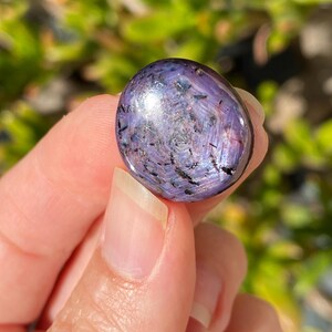 May include: A polished, oval-shaped gemstone with a swirling pattern of purple, black, and silver. The stone is held up against a blurred green background, showcasing its iridescent surface and unique color variations.