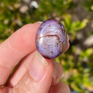 May include: A polished, oval-shaped gemstone with a lavender base color, featuring streaks of brown and flecks of black. The stone is held between fingers, with a blurred green background. The gemstone's surface reflects light, showcasing its unique patterns and textures.