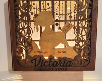 Op de afbeelding: Houten nachtlampje met silhouet van een lezend kind. De donkerbruine lijst heeft ingewikkelde uitgesneden patronen. Het licht toont de naam "Victoria", geboortegewicht 3000 g, lengte 47 cm en de datum 03.03.2009.