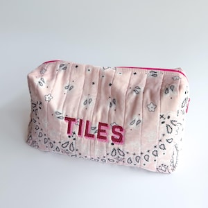 May include: A rectangular quilted pouch in light pink featuring a black paisley print pattern. The word TILES is embroidered in dark pink thread on the front. The pouch has a bright pink zipper closure along the top edge and a structured shape.