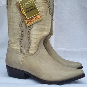 May include: A pair of light brown leather cowboy boots with a white fabric shaft. The boots have a pointed toe and a small heel. The boots have a label on the side that says "Sancho Western Boots".