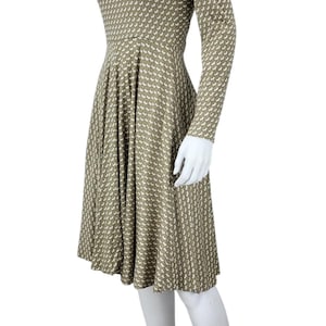 May include: A knee-length dress with a high neckline and long sleeves. The dress features a repeating pattern of small white shapes on a muted olive green background. The dress is displayed on a mannequin, paired with beige high-heeled sandals.