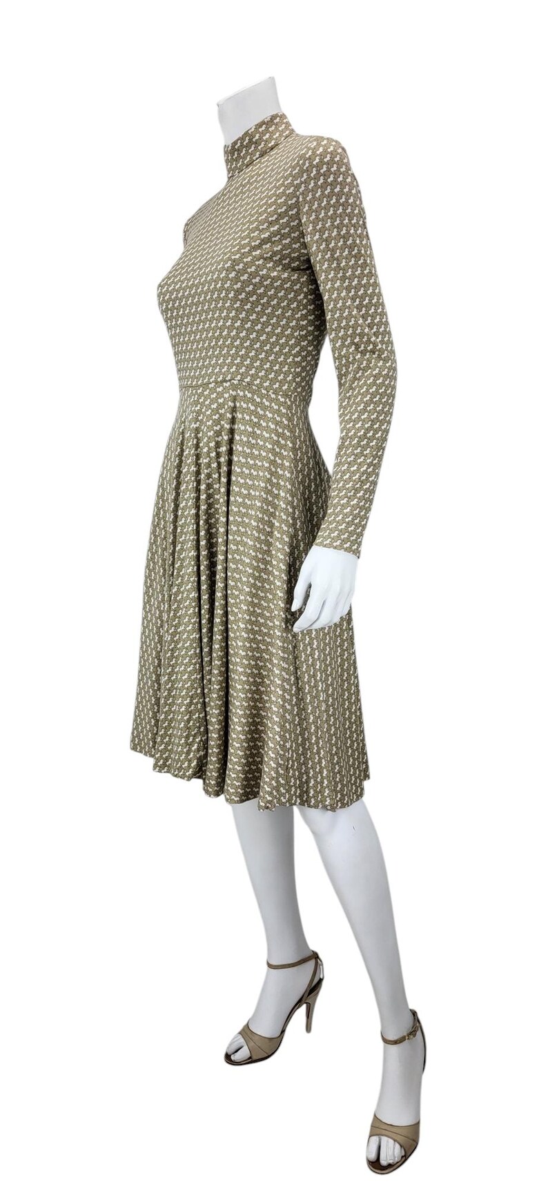 May include: A knee-length dress with a high neckline and long sleeves. The dress features a repeating pattern of small white shapes on a muted olive green background. The dress is displayed on a mannequin, paired with beige high-heeled sandals.