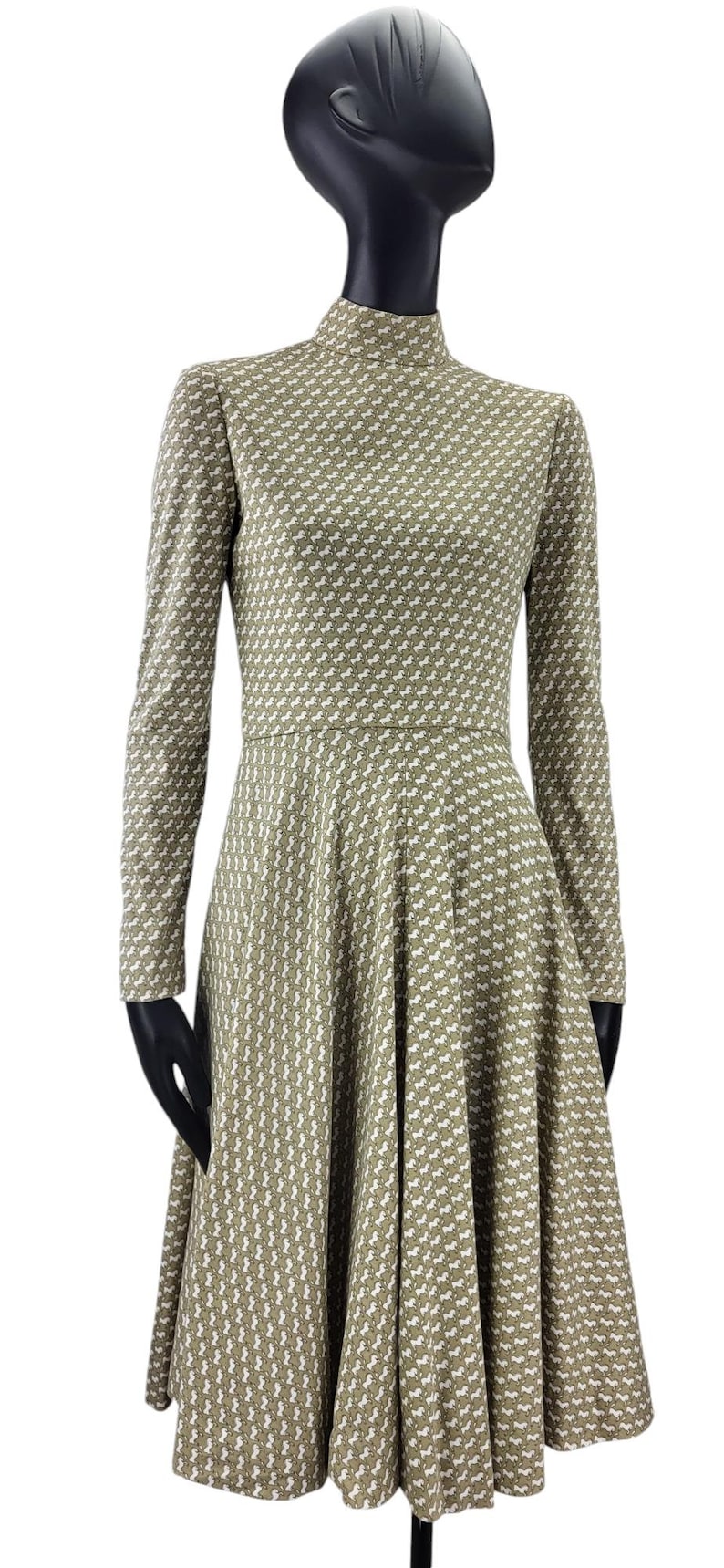 May include: A long-sleeved, olive green dress with a high neckline and a flared skirt. The dress features a repeating white pattern. The dress is displayed on a mannequin.