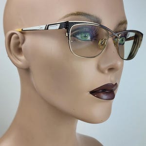 May include: Gold-rimmed eyeglasses with a black and white patterned accent on the temples. The glasses have a rectangular shape and are set against a neutral background. The lenses are tinted.