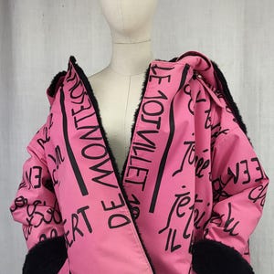 Può includere: Una giacca rosa con cappuccio con finiture in finta pelliccia nera e testo nero. La giacca è esposta su un manichino. Il testo sembra essere in un carattere di scrittura. La giacca ha una vestibilità ampia.