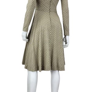 May include: A long-sleeved, knee-length dress on a mannequin. The dress features a high neckline and a fitted bodice that flares into a skirt. The fabric is a tan color with a repeating white pattern. The mannequin is wearing beige high-heeled sandals.