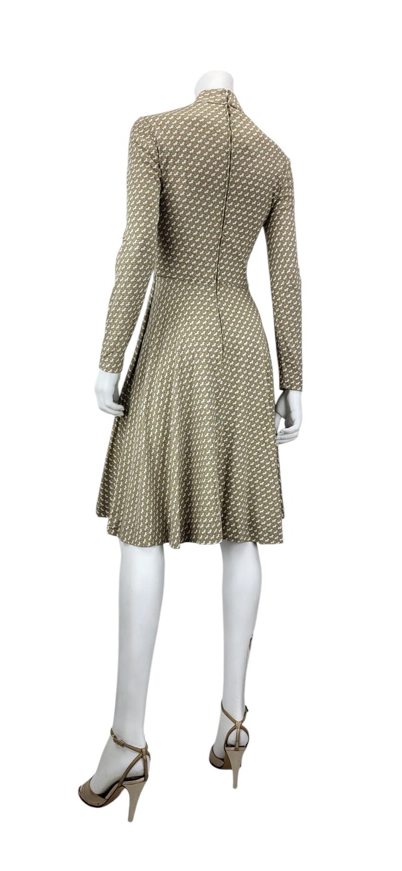 May include: A long-sleeved, knee-length dress on a mannequin. The dress features a high neckline and a fitted bodice that flares into a skirt. The fabric is a tan color with a repeating white pattern. The mannequin is wearing beige high-heeled sandals.