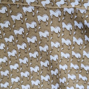 May include: Tan fabric with a repeating pattern of white horses. The horses are depicted in a running pose, creating a dynamic and lively design. The fabric appears to be a soft material, suitable for clothing or home decor.