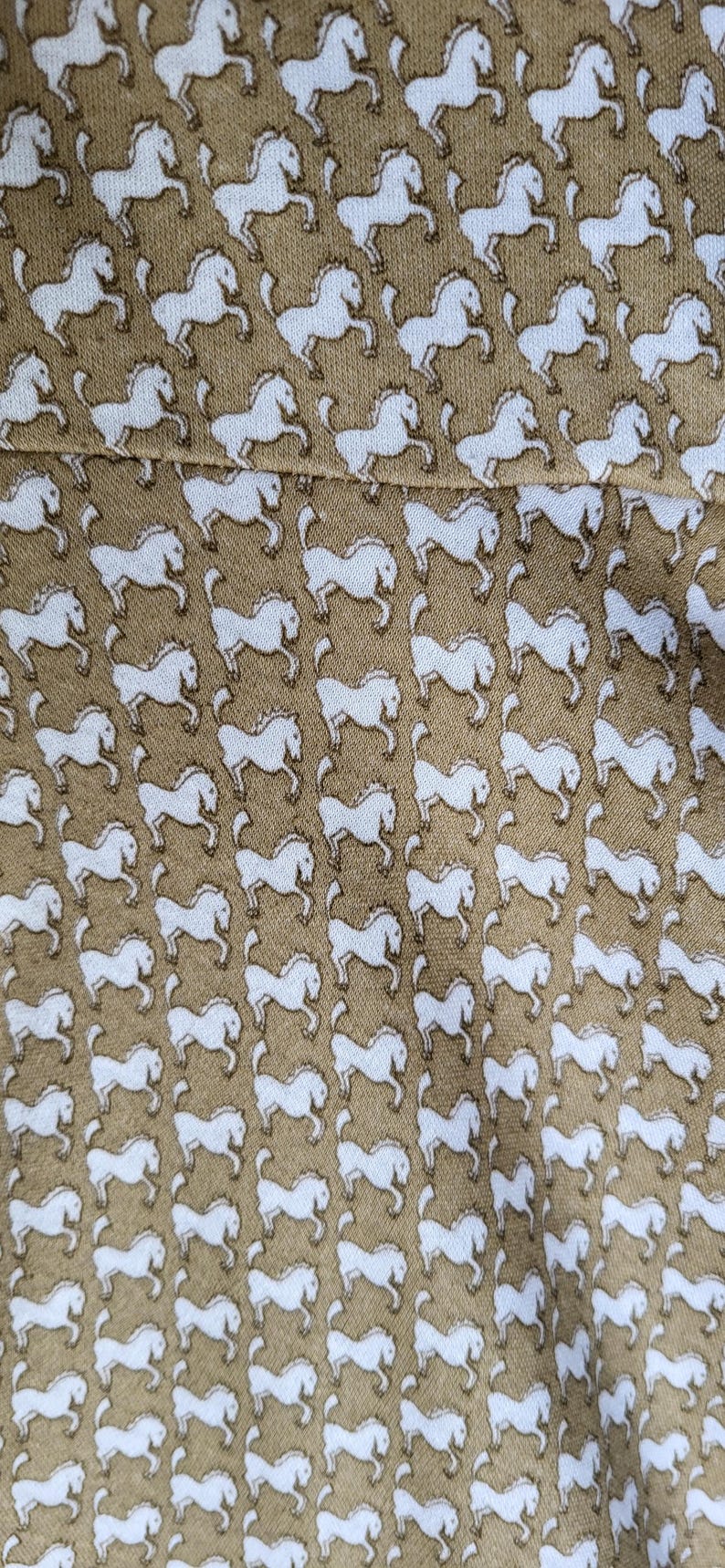 May include: Tan fabric with a repeating pattern of white horses. The horses are depicted in a running pose, creating a dynamic and lively design. The fabric appears to be a soft material, suitable for clothing or home decor.