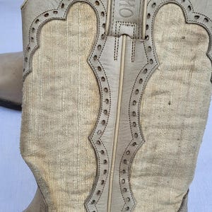 May include: A pair of light brown cowboy boots with a textured fabric upper and a leather sole. The boots have a pointed toe and a small heel. The boots are decorated with a stitching detail around the top of the boot and a leather strap with the word "SANCHO" embossed on it.