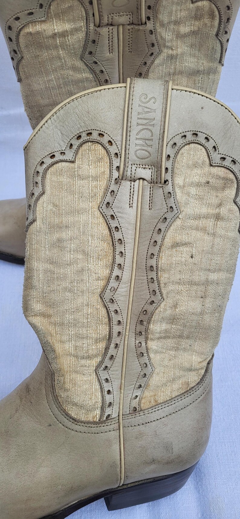 May include: A pair of light brown cowboy boots with a textured fabric upper and a leather sole. The boots have a pointed toe and a small heel. The boots are decorated with a stitching detail around the top of the boot and a leather strap with the word "SANCHO" embossed on it.