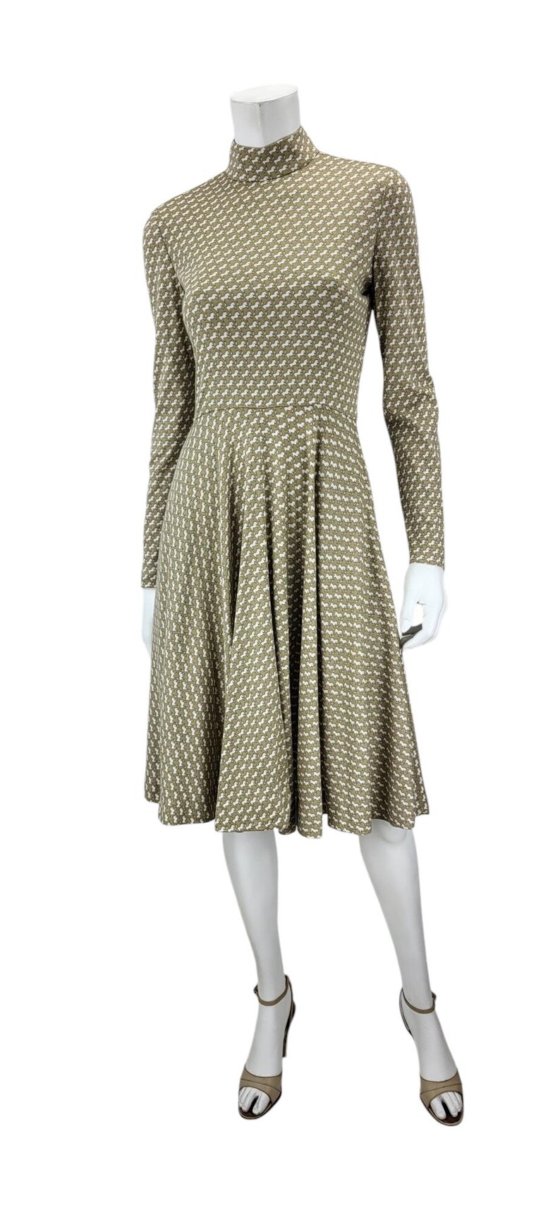 May include: A long-sleeved dress with a high neckline, featuring a repeating white and olive green pattern. The dress has a fitted bodice and a flared skirt, displayed on a mannequin. The mannequin is wearing beige heels.