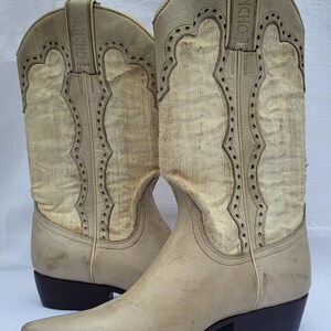 May include: A pair of light beige cowboy boots with a pointed toe and a small heel. The boots have a decorative stitching pattern and the brand name "SANCHO" is embossed on the side.