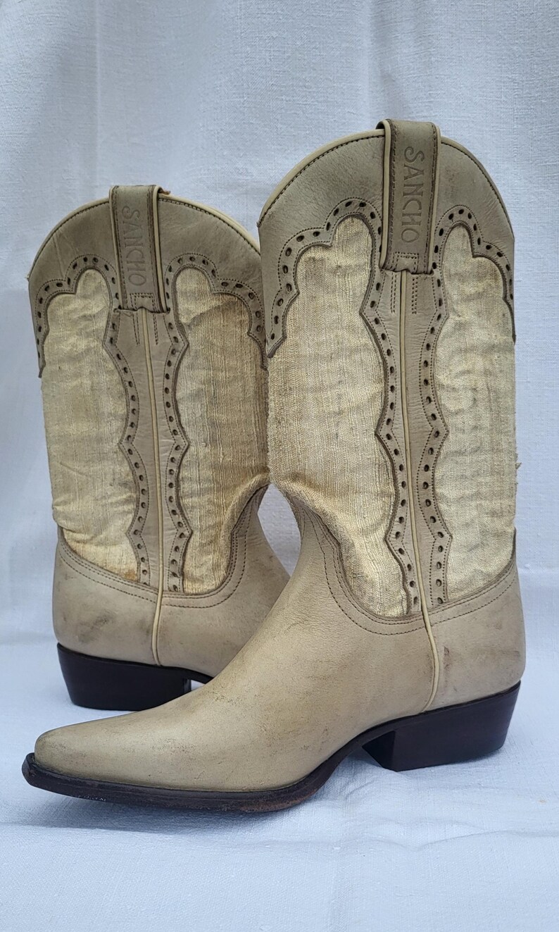 May include: A pair of light beige cowboy boots with a pointed toe and a small heel. The boots have a decorative stitching pattern and the brand name "SANCHO" is embossed on the side.