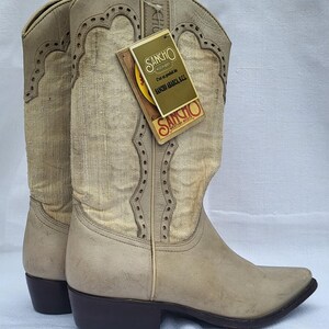 May include: A pair of off-white cowboy boots with a pointed toe and a brown heel. The boots have a decorative stitching pattern and a fabric panel on the shaft. The boots are labeled "Sancho America Kal" on a tag.