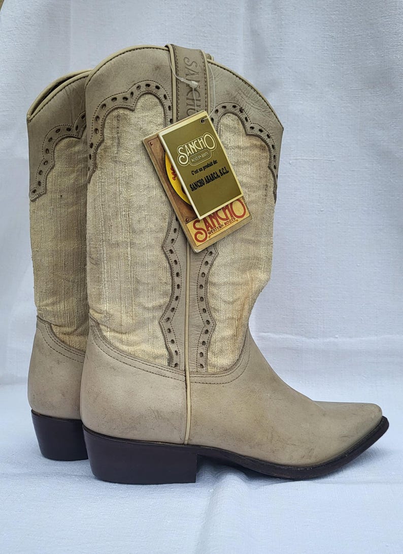 May include: A pair of off-white cowboy boots with a pointed toe and a brown heel. The boots have a decorative stitching pattern and a fabric panel on the shaft. The boots are labeled "Sancho America Kal" on a tag.