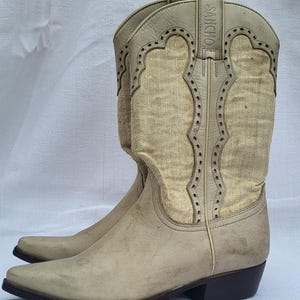 May include: A pair of off-white cowboy boots with a pointed toe and a small heel. The boots have a decorative stitching pattern and the brand name "SANCHO" is printed on the side.