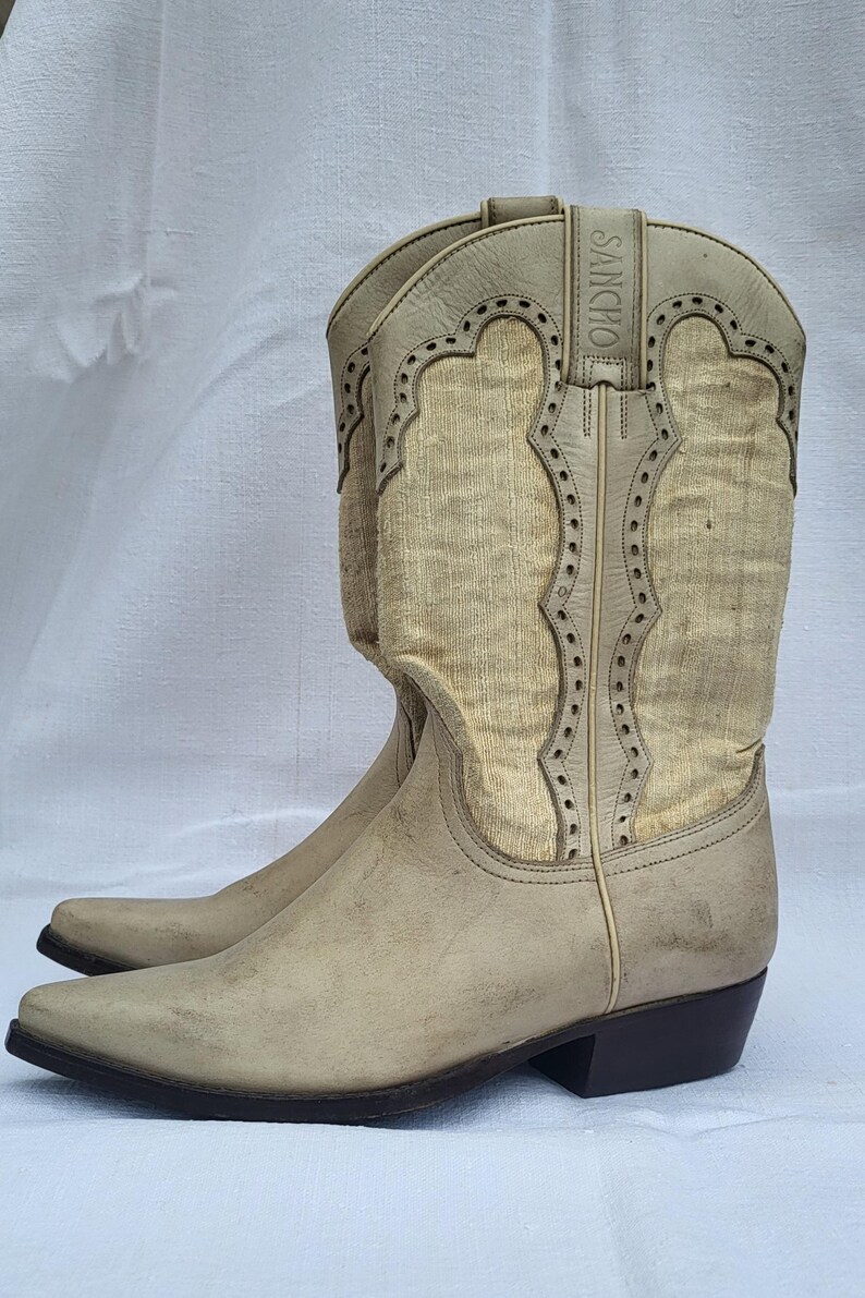 May include: A pair of off-white cowboy boots with a pointed toe and a small heel. The boots have a decorative stitching pattern and the brand name "SANCHO" is printed on the side.
