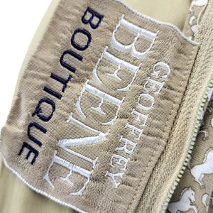 May include: Close-up of a beige fabric with a white horse pattern and a zipper. A label with the text "BEAUVE" and "GEOFFREY BOUTIQUE" is stitched on the fabric. The zipper is beige.