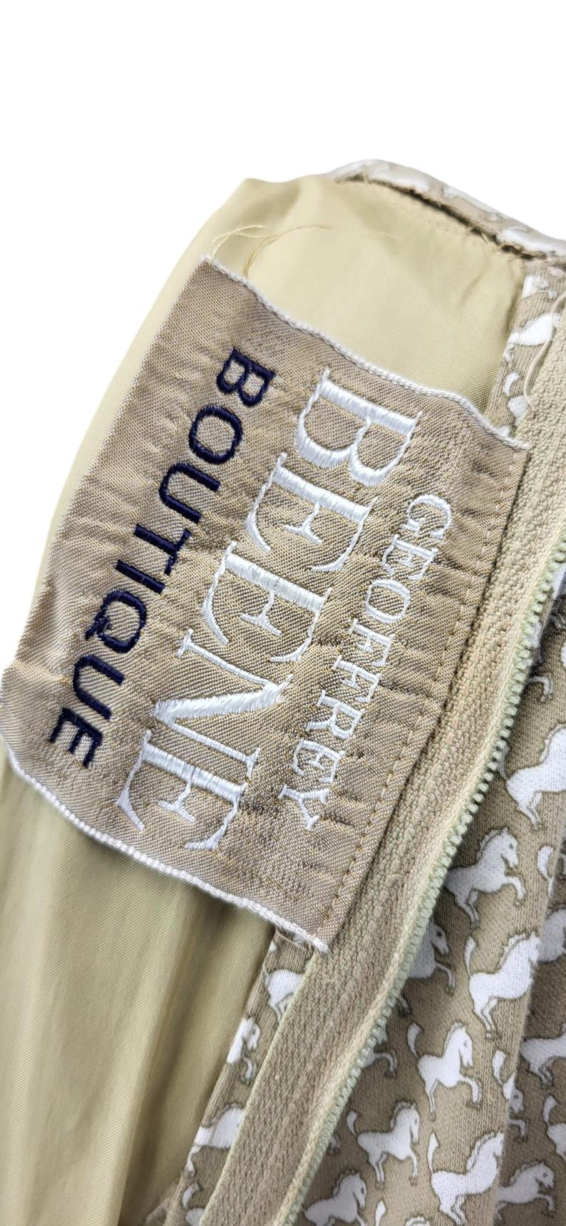 May include: Close-up of a beige fabric with a white horse pattern and a zipper. A label with the text "BEAUVE" and "GEOFFREY BOUTIQUE" is stitched on the fabric. The zipper is beige.
