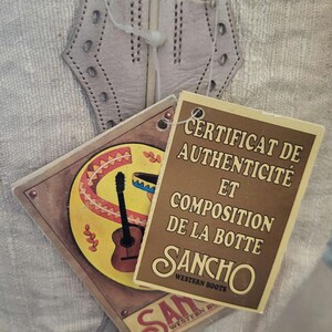 May include: A close-up of a white leather cowboy boot with a brown tag that reads "Certificat de Authenticit&eacute; et Composition de la Botte Sancho Western Boots".