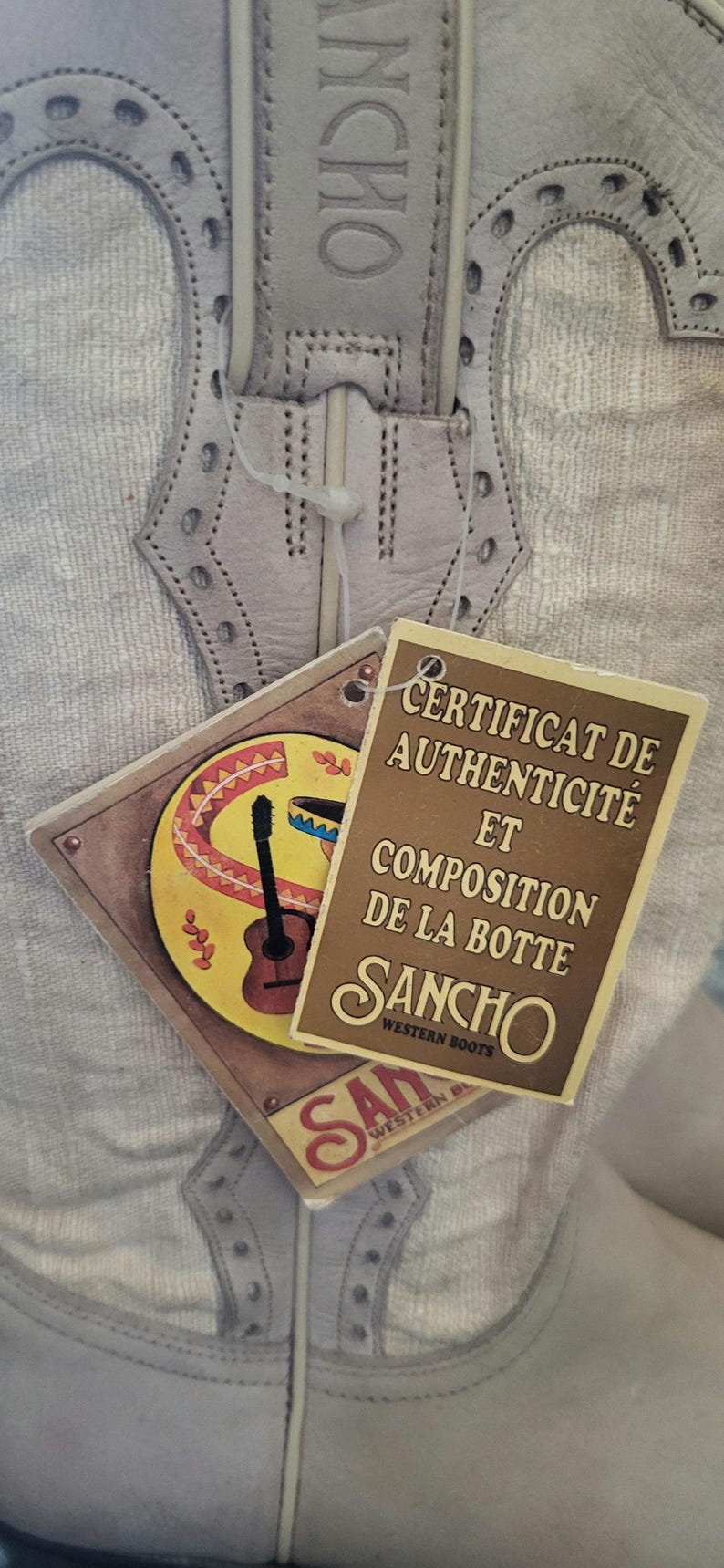 May include: A close-up of a white leather cowboy boot with a brown tag that reads "Certificat de Authenticit&eacute; et Composition de la Botte Sancho Western Boots".