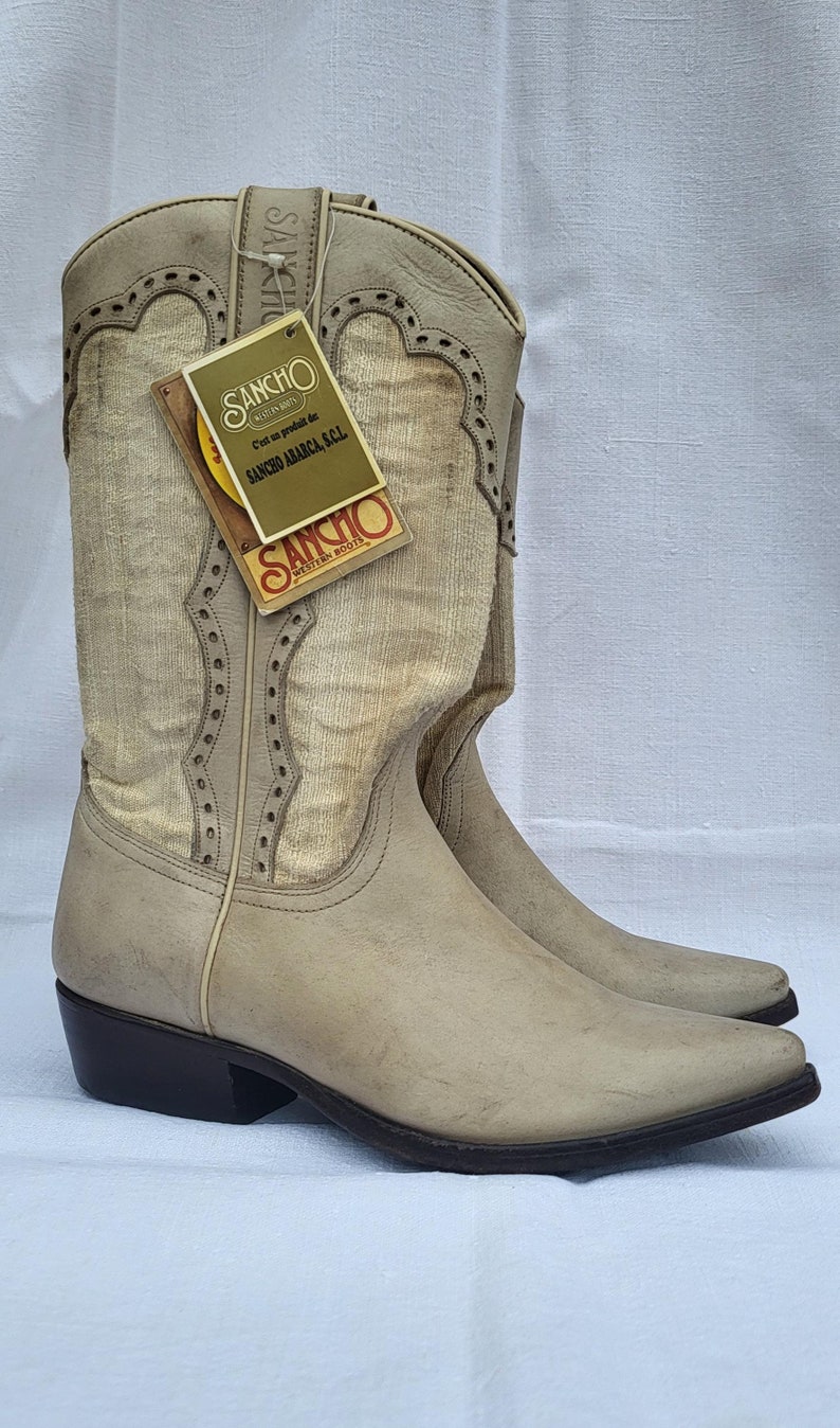 May include: A pair of light brown leather cowboy boots with a white fabric shaft. The boots have a pointed toe and a small heel. The boots have a label on the side that says "Sancho Western Boots".