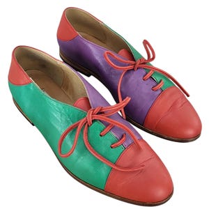 以下が含まれることがあります： 赤、緑、紫のパネルが付いたカラフルな革靴のペア。靴には靴ひもがあり、丸みを帯びたつま先です。