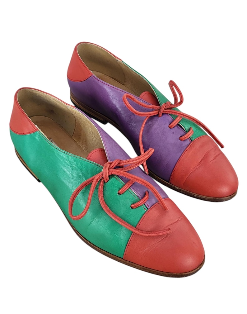 以下が含まれることがあります： 赤、緑、紫のパネルが付いたカラフルな革靴のペア。靴には靴ひもがあり、丸みを帯びたつま先です。