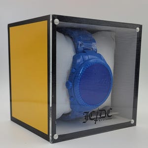 May include: A blue wristwatch with a matching band is displayed inside a clear acrylic box with a yellow and black accent. The watch is on a white pillow. The box has the text "JC/DC" on a tag.