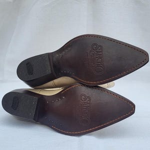 May include: A pair of beige leather cowboy boots with brown leather soles. The soles are marked with the text "Sancho Western Collection" and the size "36".