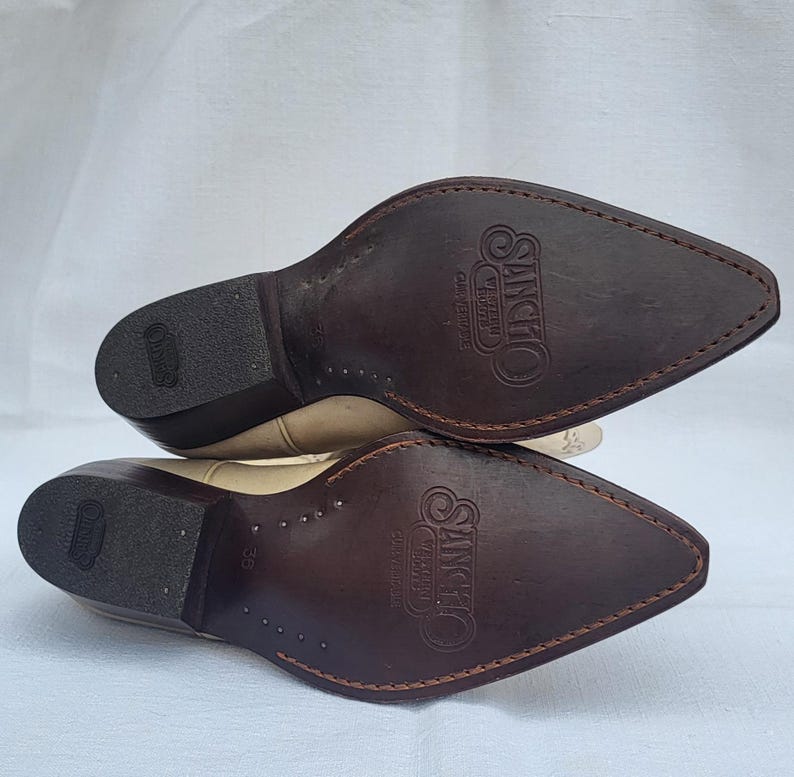 May include: A pair of beige leather cowboy boots with brown leather soles. The soles are marked with the text "Sancho Western Collection" and the size "36".