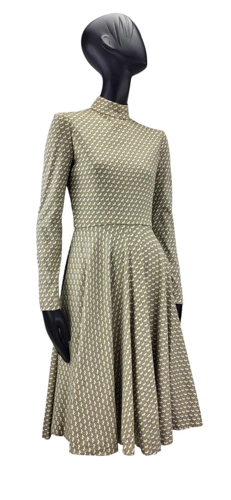 May include: A long-sleeved dress with a high neckline, displayed on a mannequin. The dress features a repeating white pattern on a muted green background. The skirt flares out from the waist.