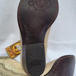 May include: A pair of light brown leather cowboy boots with a pointed toe and a decorative stitch pattern. The boots have a brown leather sole with the text "SANCHO WESTERN BOOTS" and "CUIR VERITABLE" on the heel. The size 38 is printed on the sole.