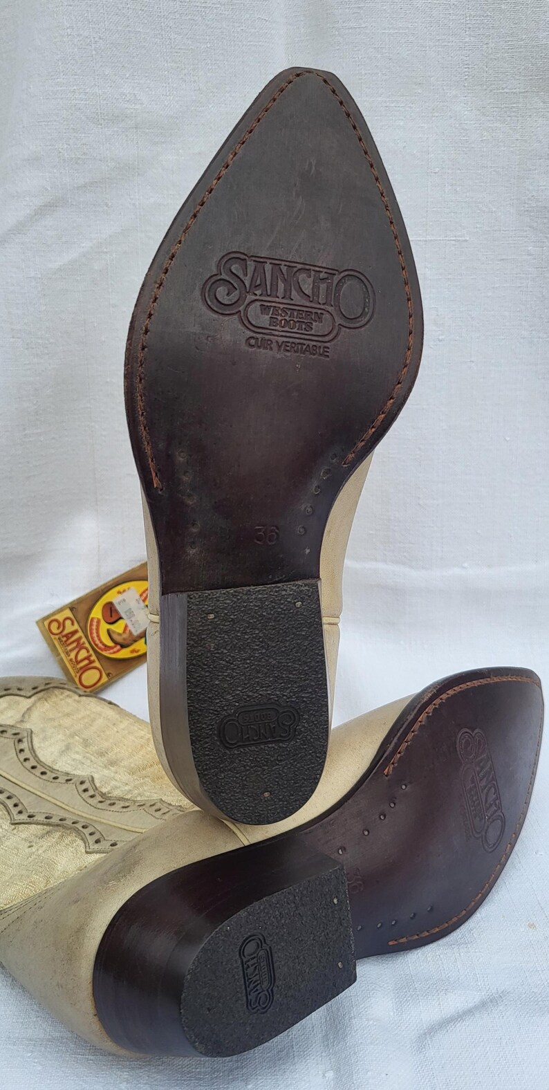May include: A pair of light brown leather cowboy boots with a pointed toe and a decorative stitch pattern. The boots have a brown leather sole with the text "SANCHO WESTERN BOOTS" and "CUIR VERITABLE" on the heel. The size 38 is printed on the sole.