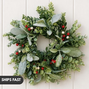May include: A circular wreath composed of green eucalyptus leaves, soft sage-colored lamb's ear foliage, and small clusters of red and white berries. The arrangement features a mix of textures and shades of green, creating a dense, layered appearance.