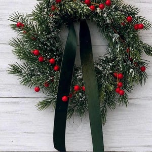 May include: A circular wreath constructed from artificial green pine branches dusted with white snow-like particles. The wreath is decorated with clusters of small, bright red berries and features two long, dark green velvet ribbons hanging down.