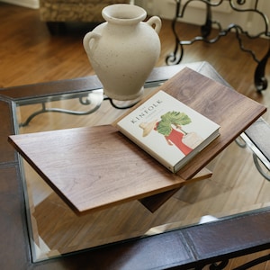May include: A wooden, two-tiered coffee table with a glass top. The top tier holds a book titled "The Kinfolk Garden" and a cream-colored vase. The table is surrounded by a brown leather frame and sits on a hardwood floor.