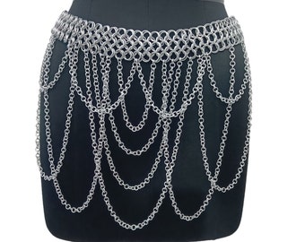 May include: Silver-colored chain belt with a layered design. The belt features a series of interconnected rings forming a wide band, with multiple strands of chains cascading downwards. The belt is displayed on a black mannequin.