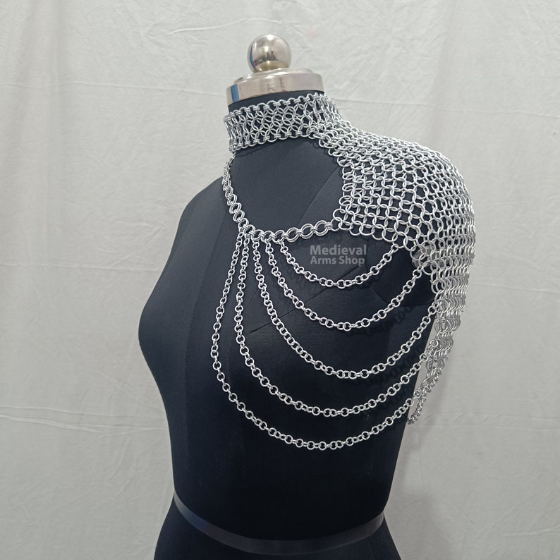 May include: Silver chainmaille shoulder armor with a matching choker necklace. The armor is made of small, interlocking metal rings and has a flowing, draped design. The necklace is a simple band of chainmaille.