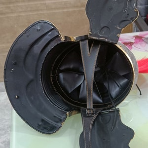 May include: A black leather helmet with a gold trim and a red plume. The helmet is open and the interior is visible. The helmet has a leather strap with a buckle.
