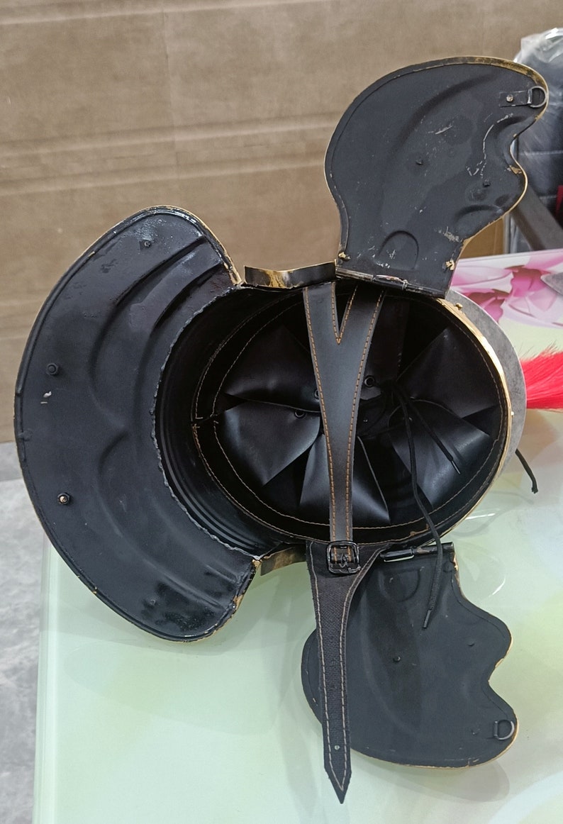 May include: A black leather helmet with a gold trim and a red plume. The helmet is open and the interior is visible. The helmet has a leather strap with a buckle.